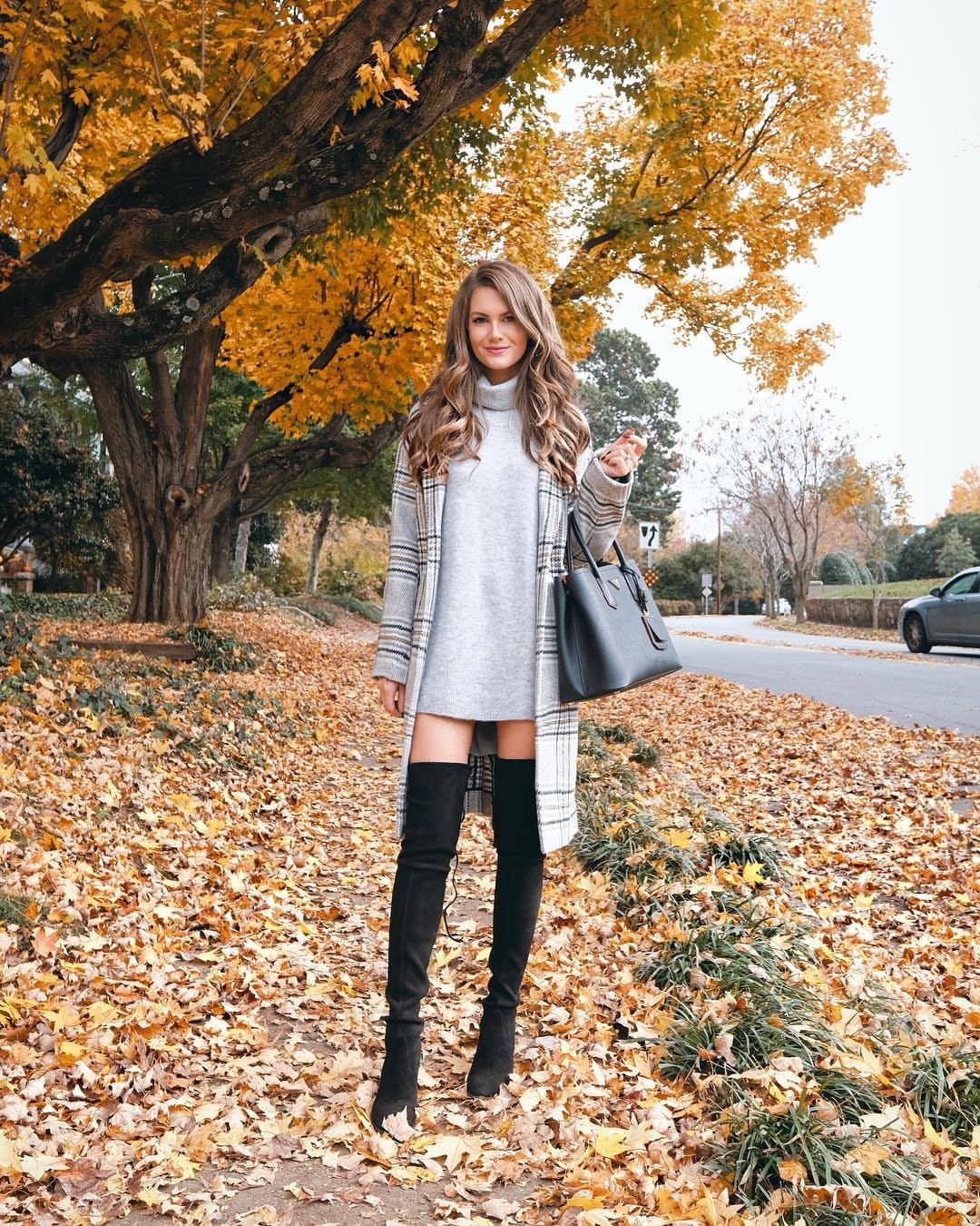 Woman wearing a grey turtleneck dress, plaid coat, and black thigh-high boots in autumn leaves.