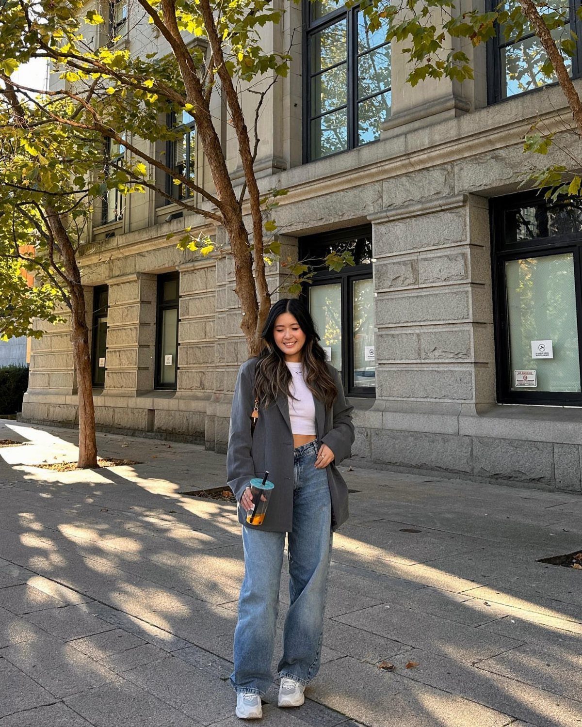 A smiling woman wearing an oversized grey blazer, white crop top, and wide-leg blue jeans.