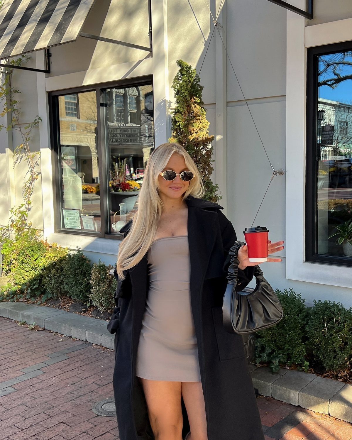 Blonde woman in a taupe mini dress and black coat holding a coffee cup outdoors.