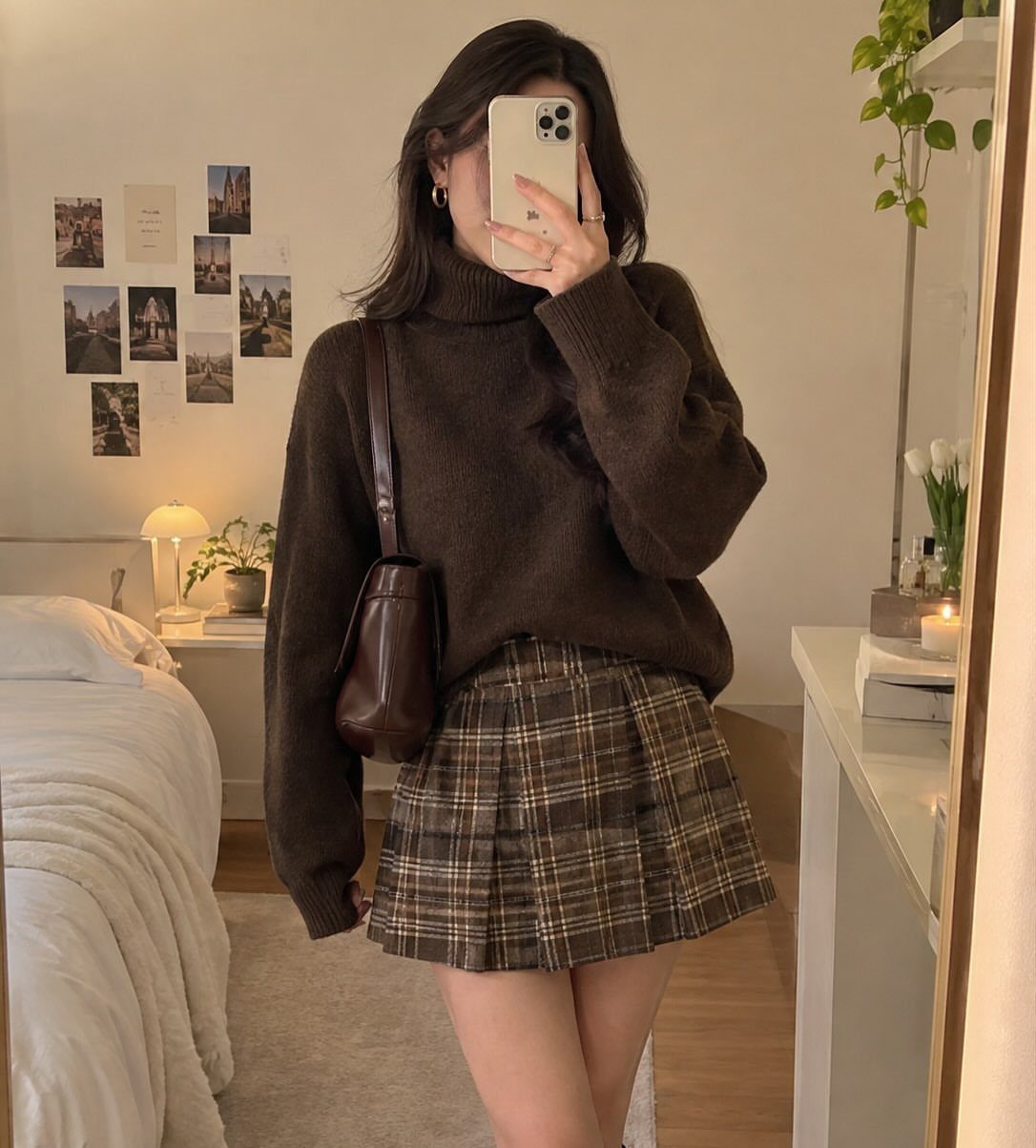 Person in a brown turtleneck sweater and plaid pleated mini skirt taking a mirror selfie.