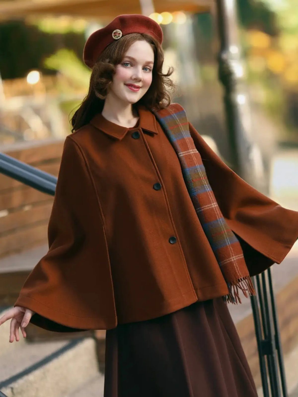 A smiling woman wearing a brown cape jacket, red beret, and plaid scarf outdoors.