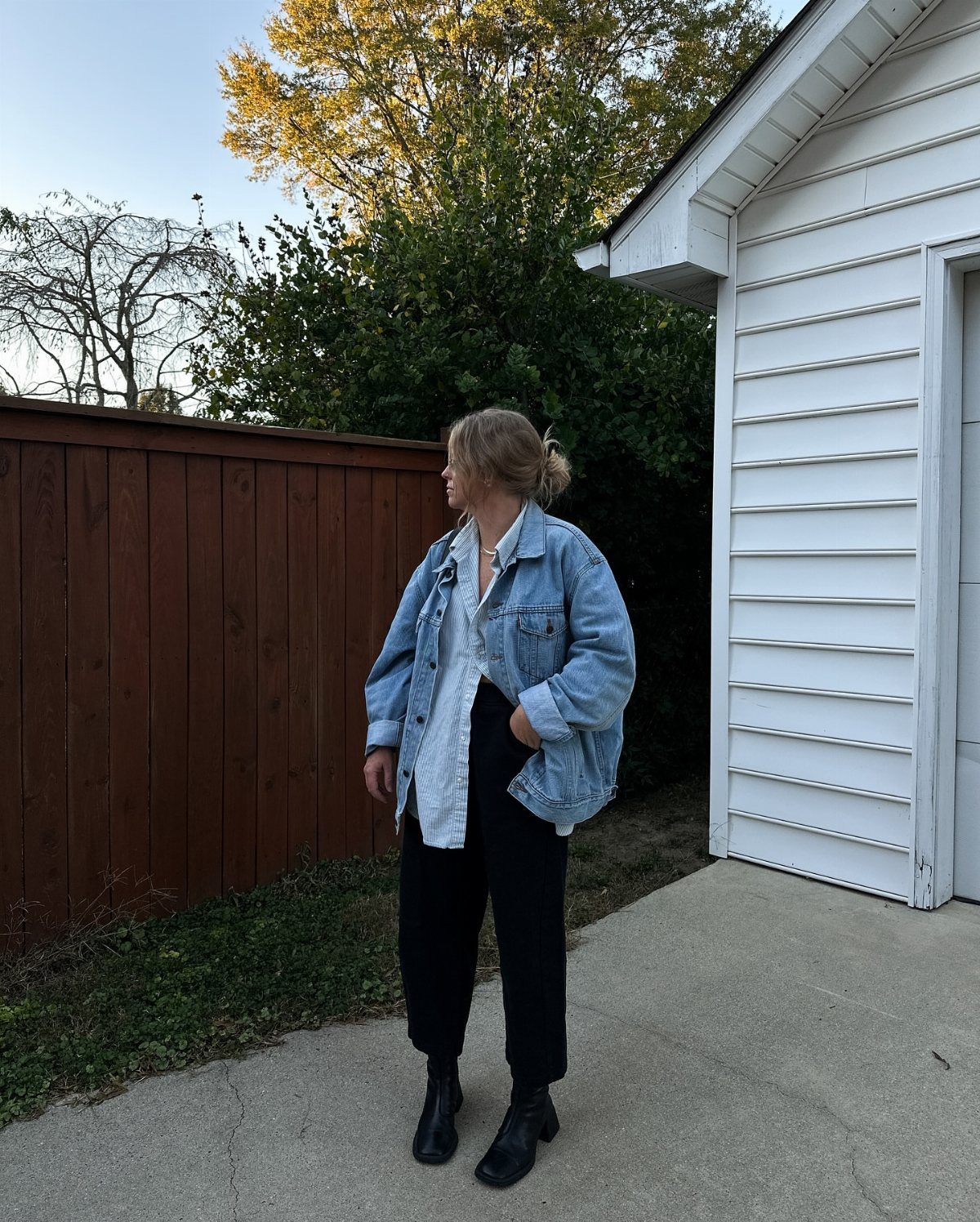 Woman wearing a denim jacket over a white button-down shirt and black trousers outdoors.