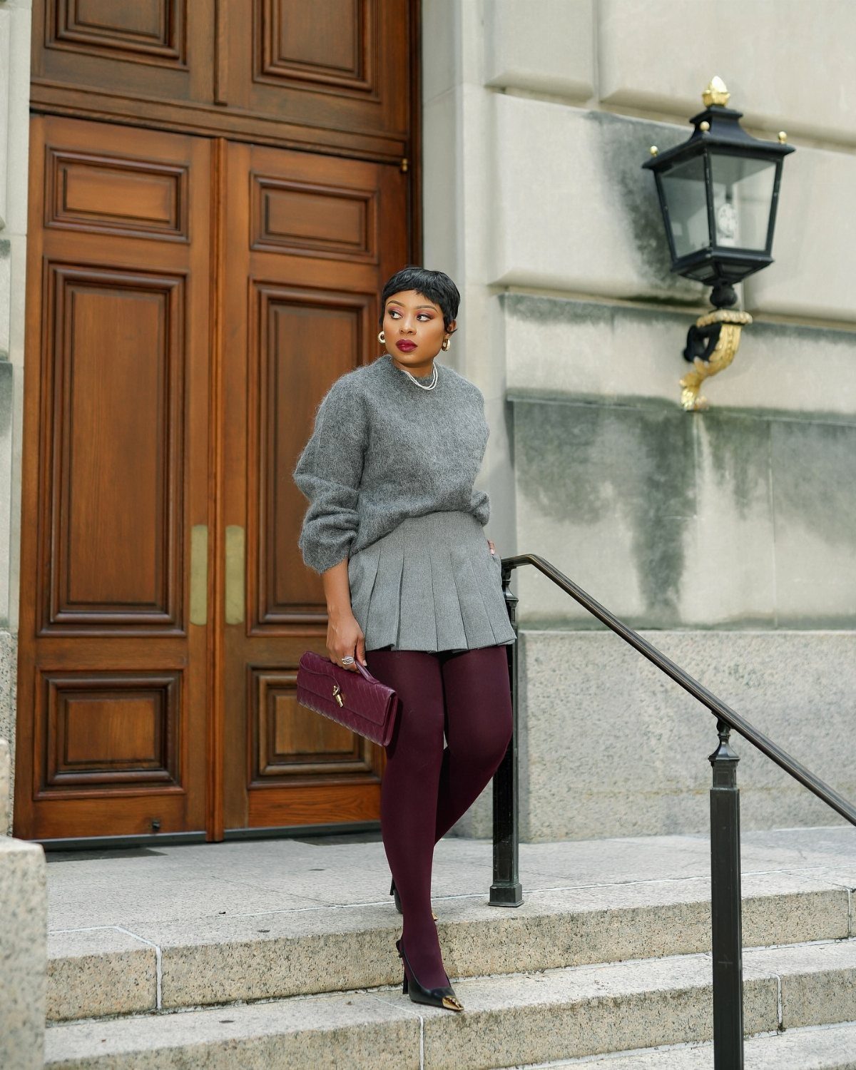 Woman in a grey sweater and pleated skirt wearing burgundy tights on stone steps.