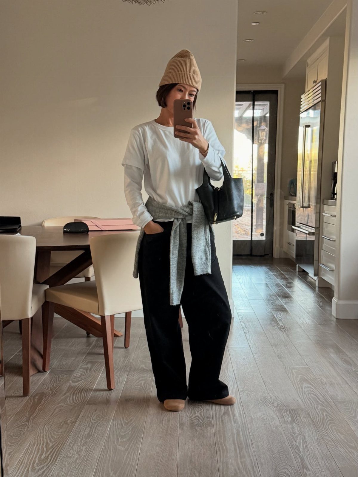 Woman in white layered tops, black trousers, and a tan beanie taking a mirror selfie.