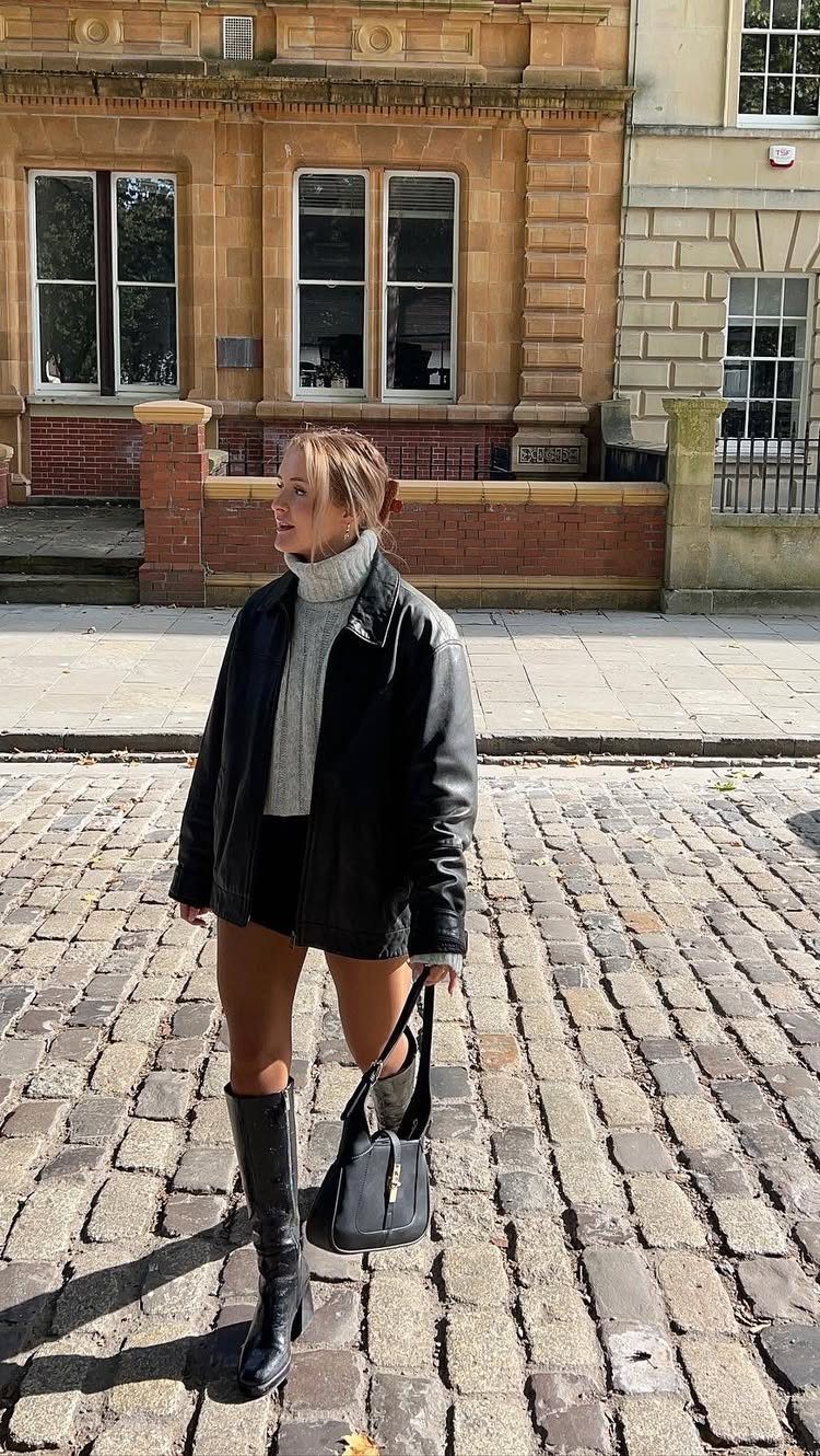 Woman in a leather jacket, white turtleneck, and black boots standing on a cobblestone street.