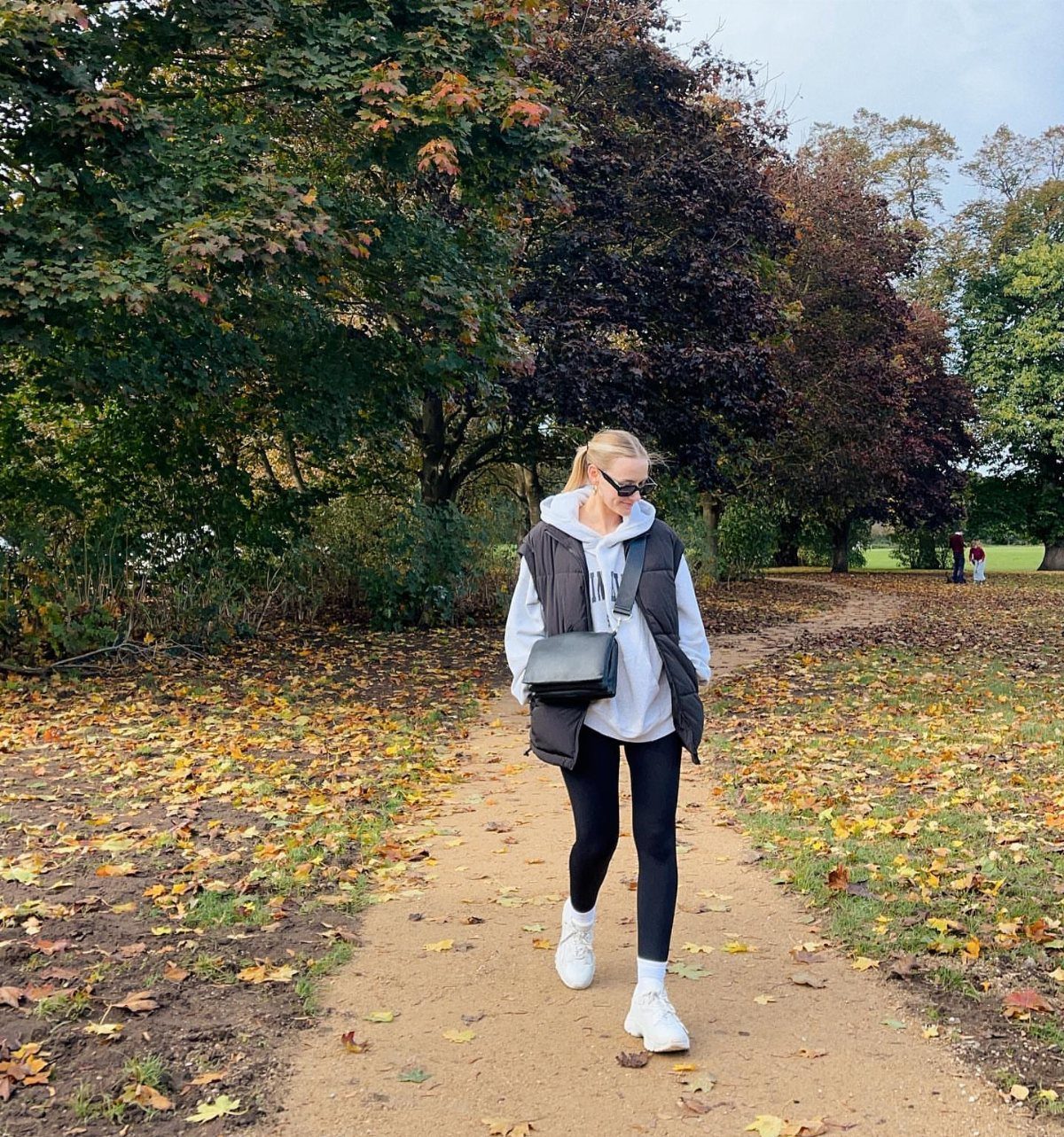 A woman wearing a white hoodie, black vest, and black leggings walks through a park.