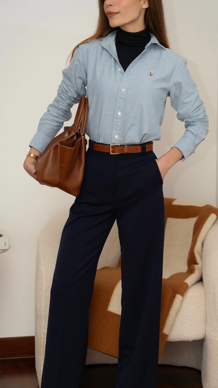 Woman wearing a light blue button-down shirt over a black ribbed turtleneck and navy trousers.