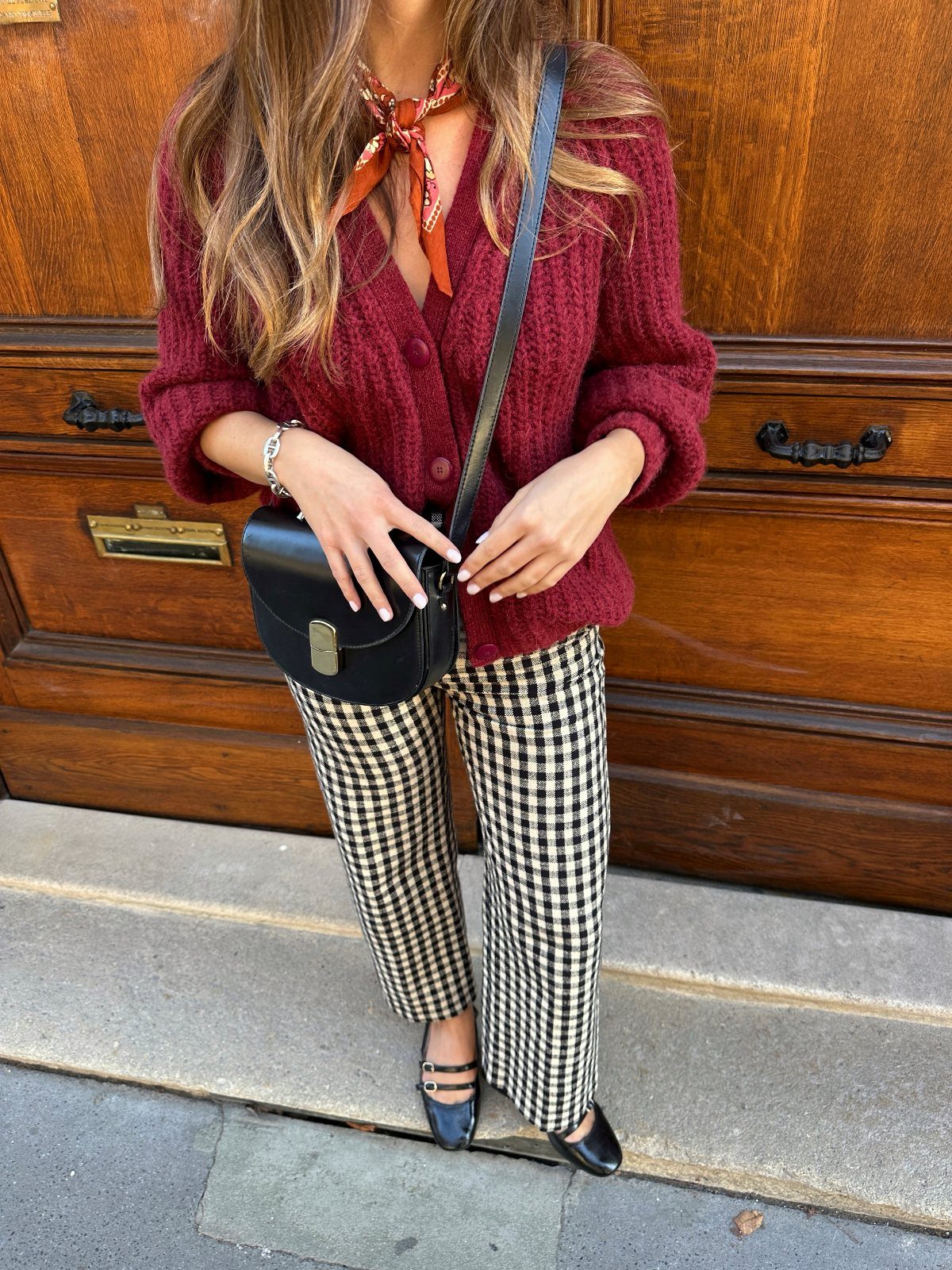 A woman wearing a burgundy cardigan and checkered trousers standing in front of a wooden door.