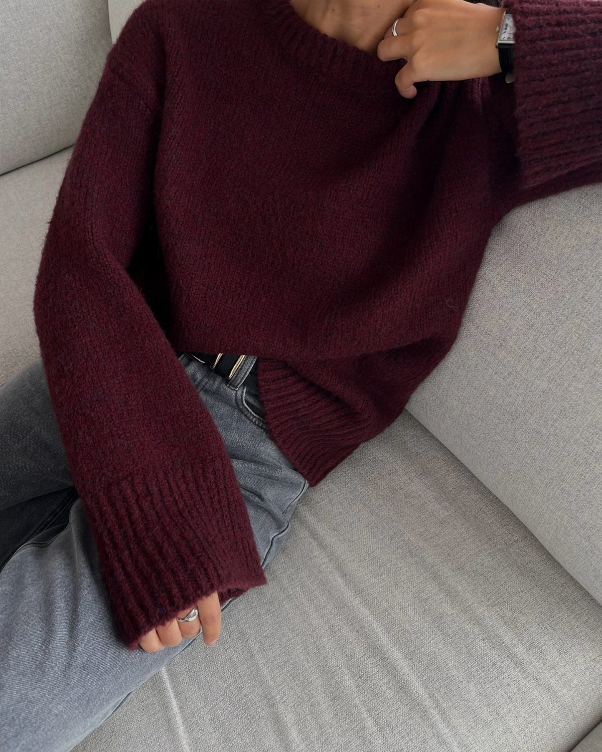 A person wearing a chunky burgundy knit sweater and grey jeans sitting on a sofa.