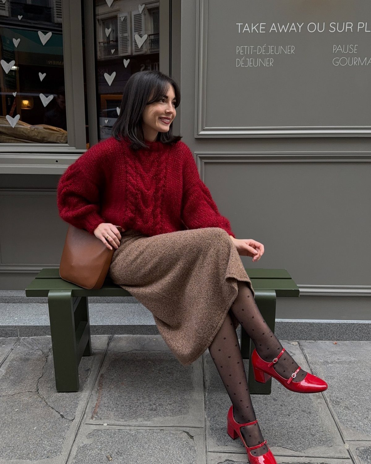 Woman in a burgundy chunky knit sweater and brown skirt sitting on a green bench.