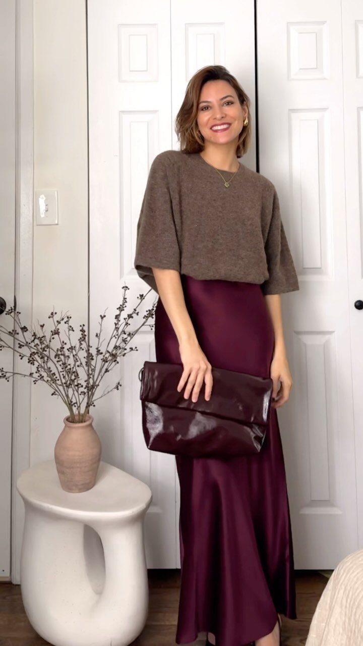 A woman wearing a brown sweater and a burgundy silk maxi skirt holding a dark clutch.