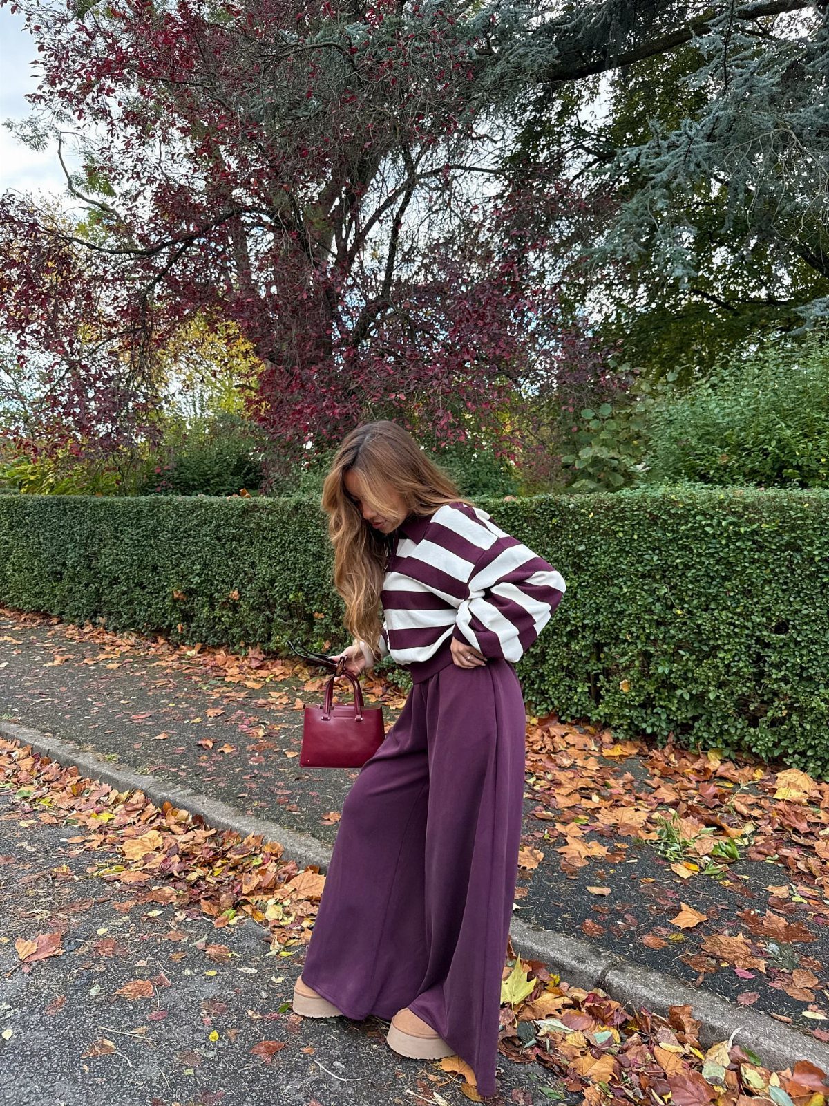 A woman wearing a striped top and burgundy wide-leg trousers standing among autumn leaves.