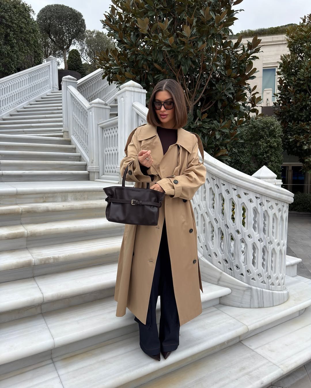 Woman in a tan trench coat and burgundy top standing on white marble stairs holding a handbag.
