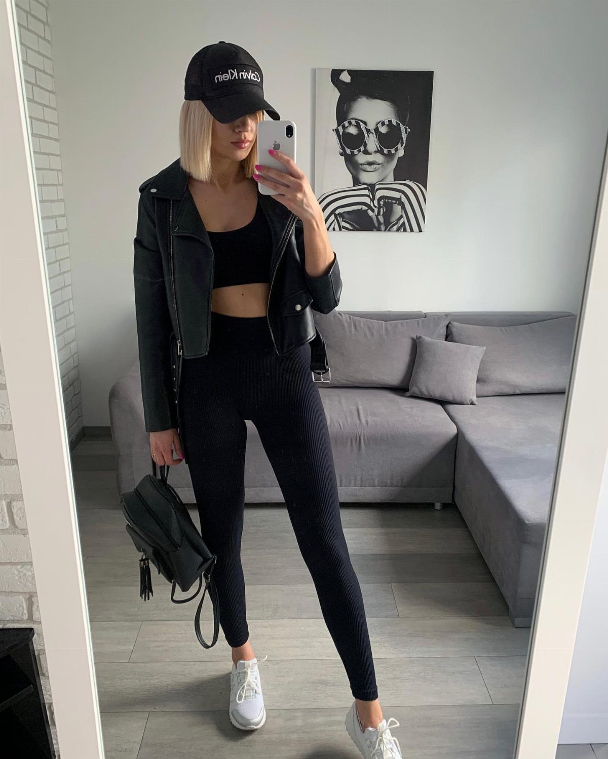 Woman in black leggings, crop top, leather jacket, and baseball cap taking a mirror selfie.