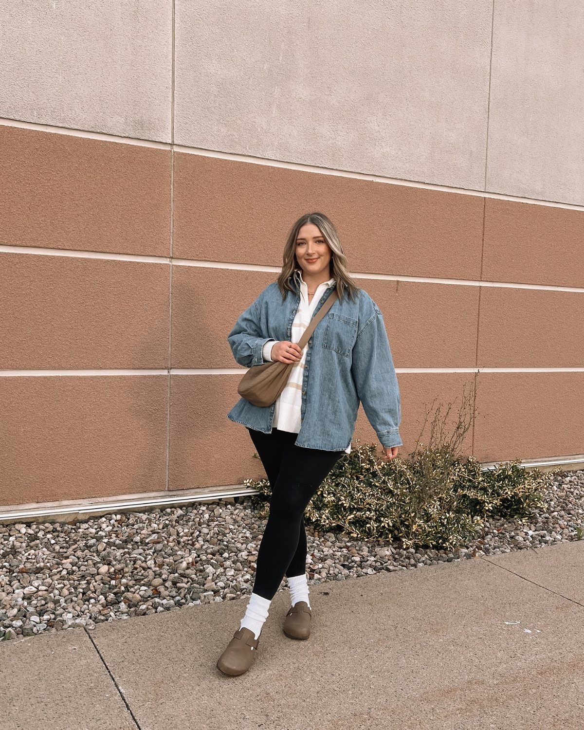 Woman wearing an oversized chambray shirt, black leggings, and brown loafers standing against a textured wall.