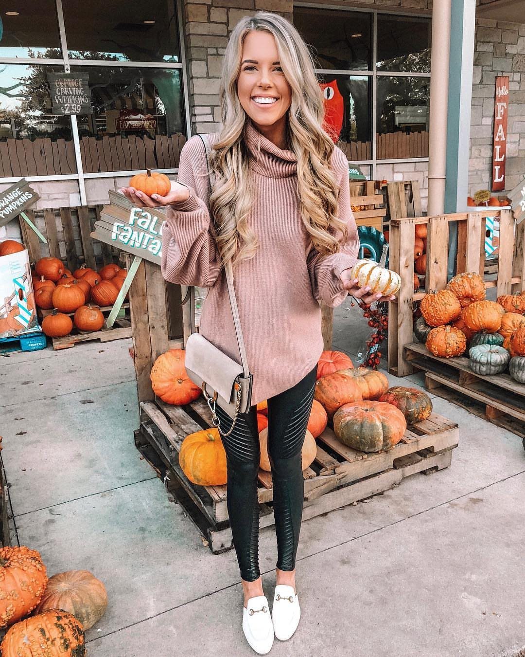 A smiling woman wearing a pink oversized sweater and black leggings holding small pumpkins outdoors.