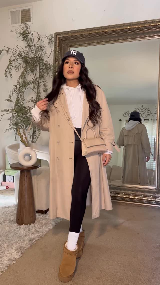 Woman posing in a long beige trench coat, white hoodie, and black leggings.