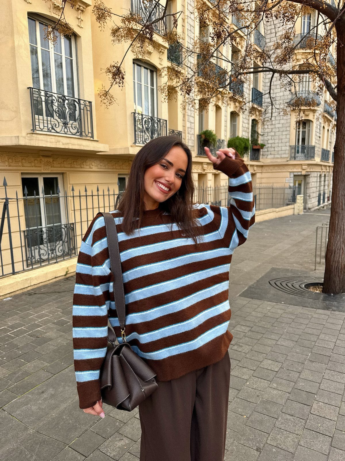 A smiling woman wearing a brown and light blue striped sweater and dark brown trousers outdoors.