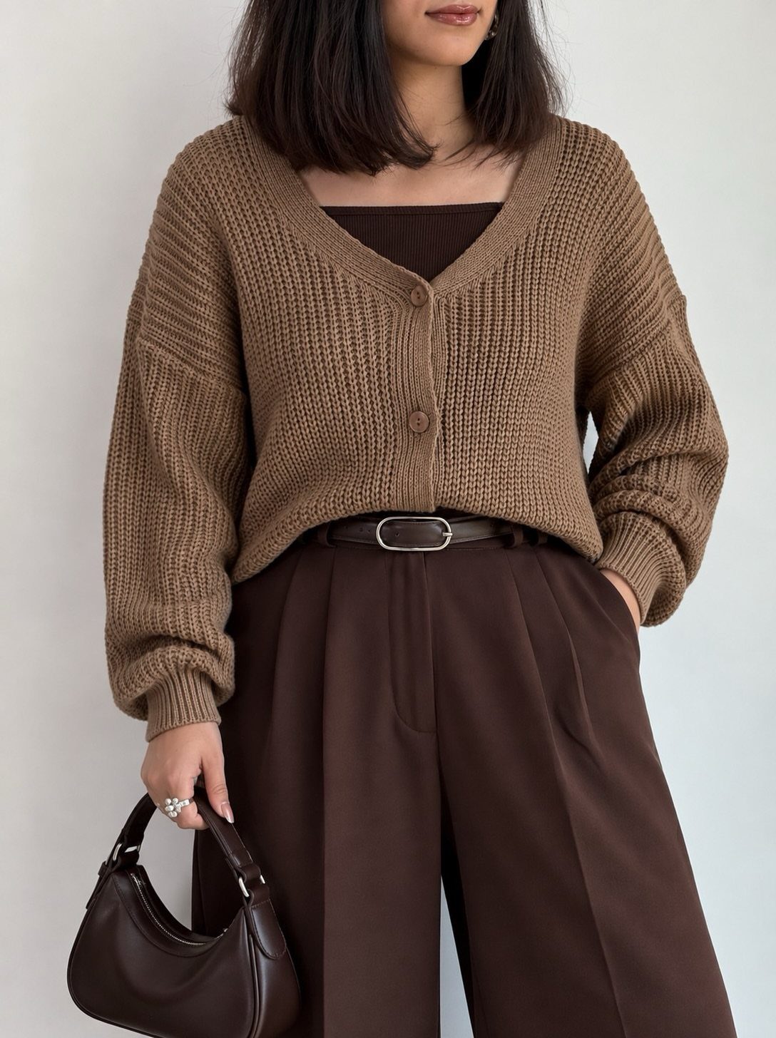 A woman wearing a brown ribbed knit cardigan and dark brown trousers holding a handbag.