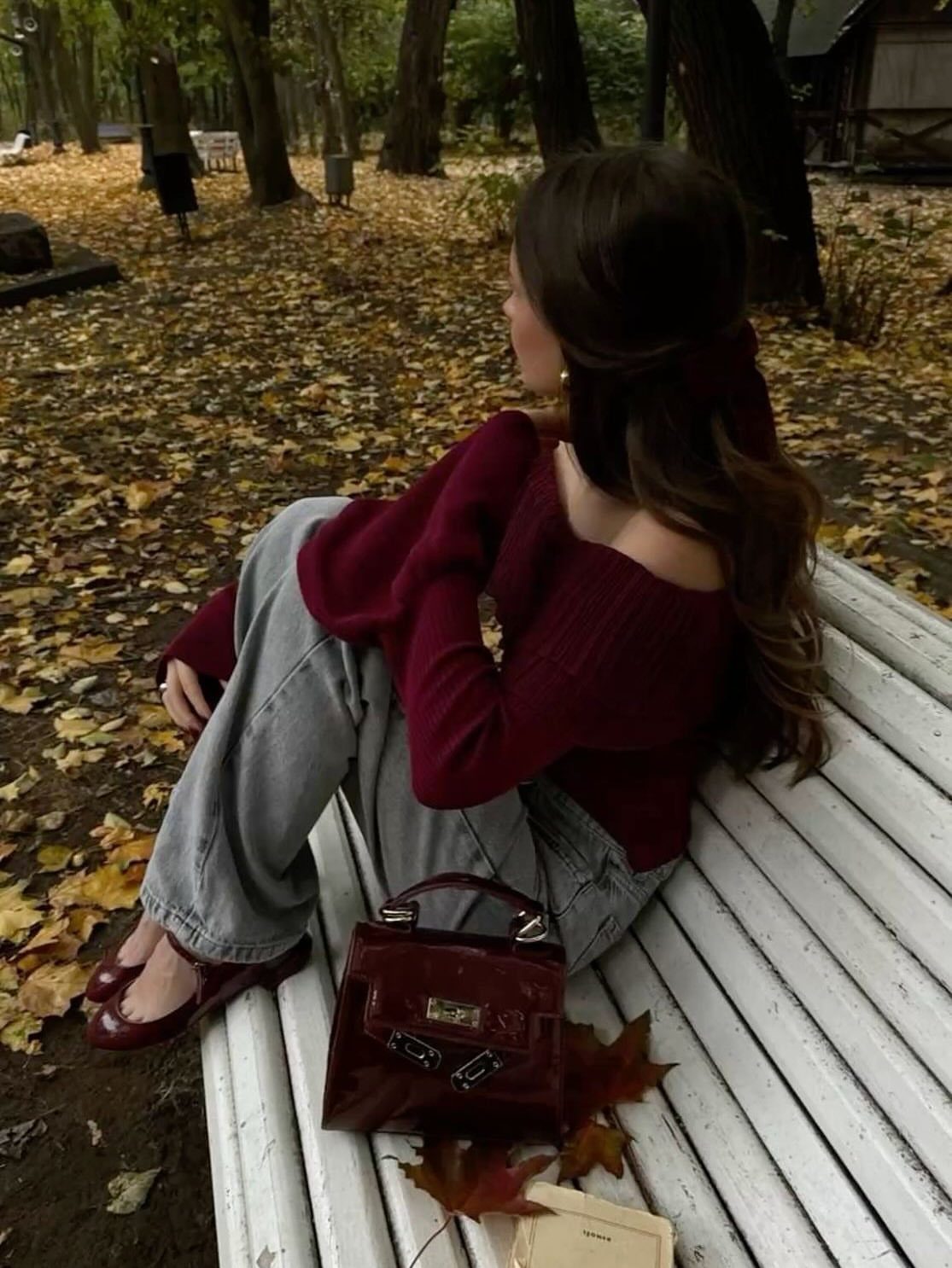 Woman in a burgundy off-the-shoulder sweater and jeans sitting on a white bench outdoors.