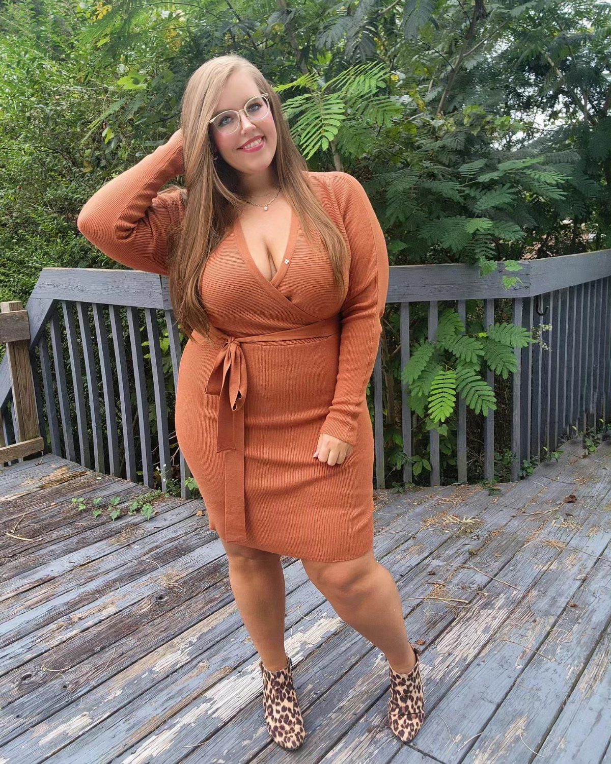 A woman in a burnt orange ribbed knit dress posing on a wooden deck outdoors.