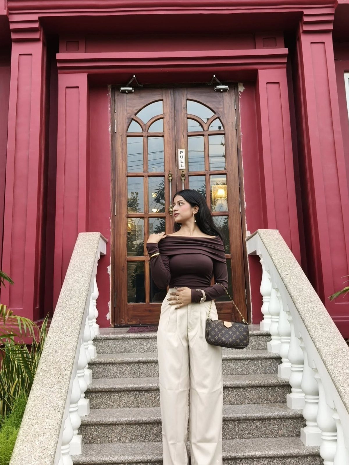 Woman in a dark brown off-the-shoulder top and cream trousers standing on outdoor stairs.