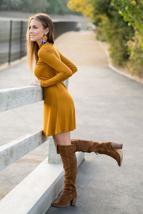 Woman in a mustard yellow mini dress and brown suede boots leaning on a white fence.
