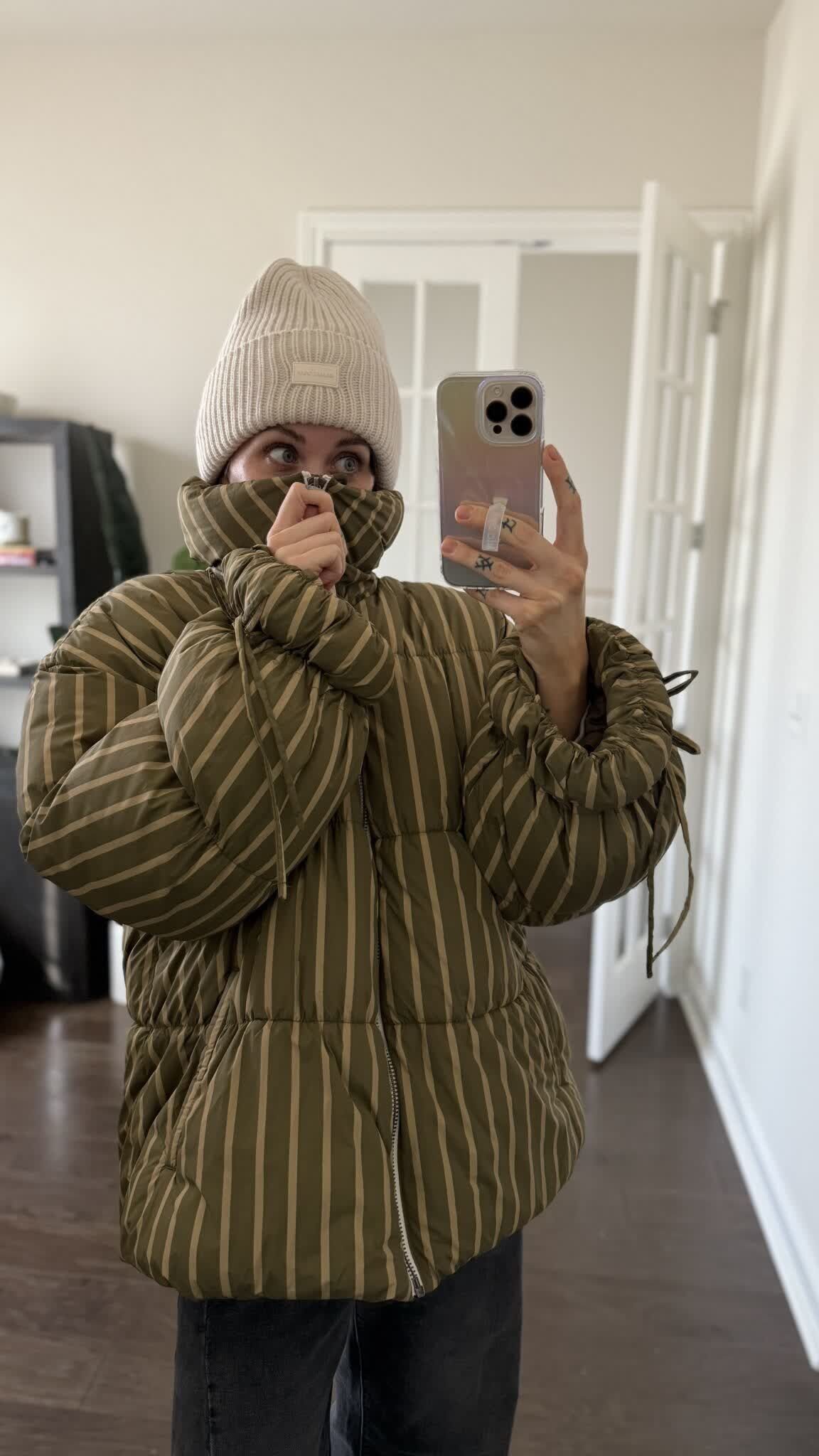 Person in a striped olive green quilted puffer jacket and beige beanie taking a mirror selfie.
