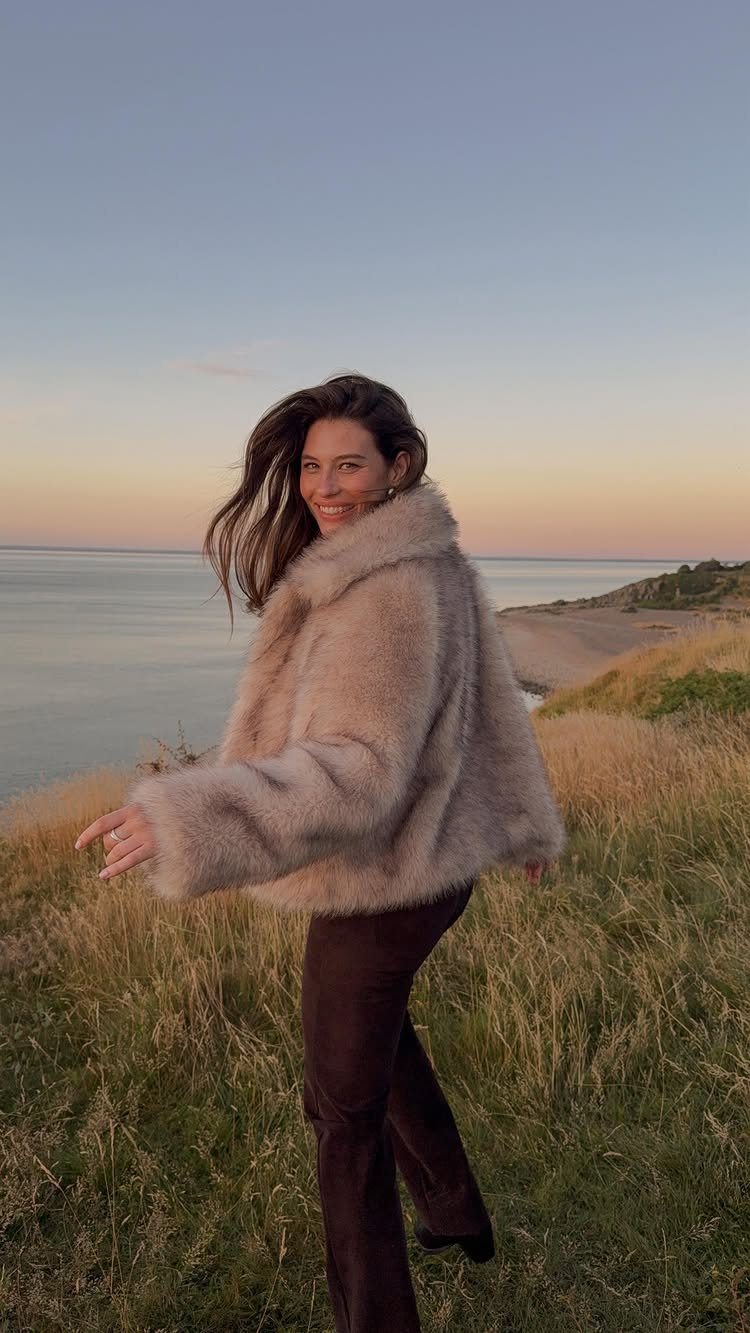 A smiling woman wearing a tan faux fur cropped jacket standing in a grassy field by the sea.