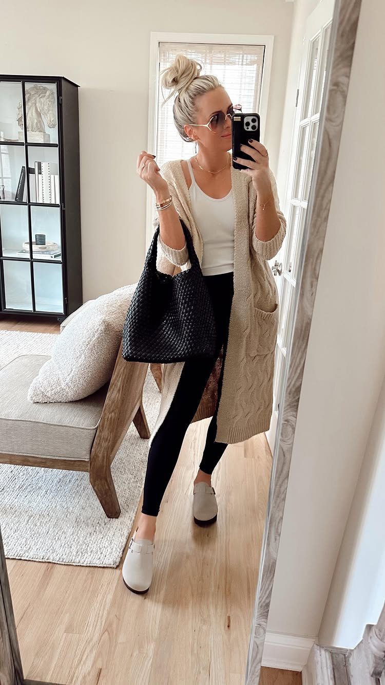 Woman in a long cream chunky knit cardigan, white tank top, and black leggings taking a selfie.