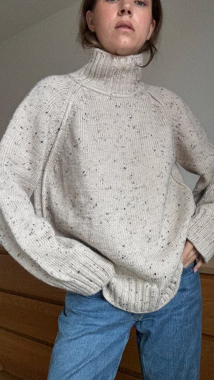 A woman wearing a thick, cream-colored speckled chunky knit turtleneck sweater and blue jeans.