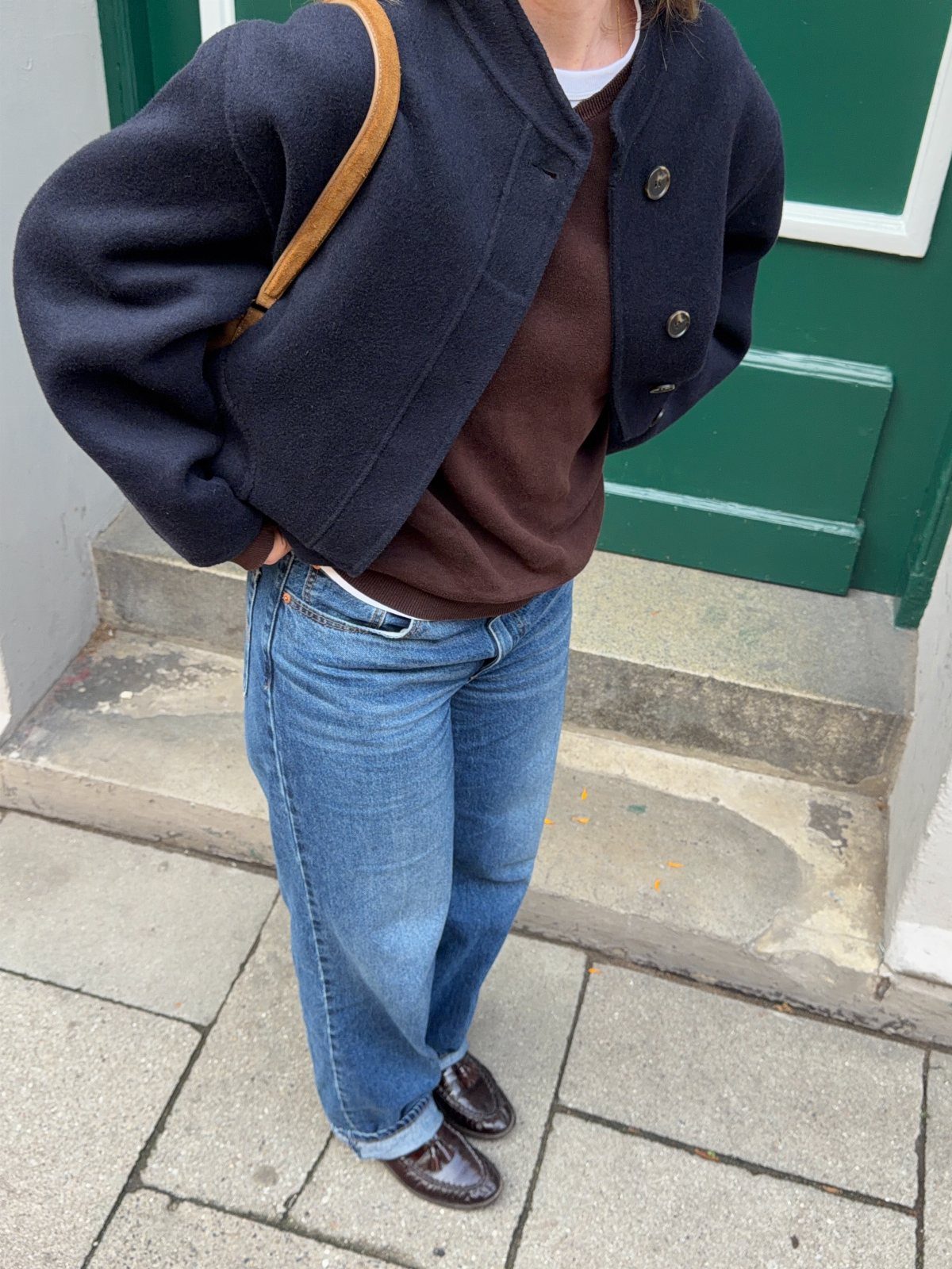 A woman wearing a navy jacket, brown sweater, blue jeans, and brown loafers standing outdoors.