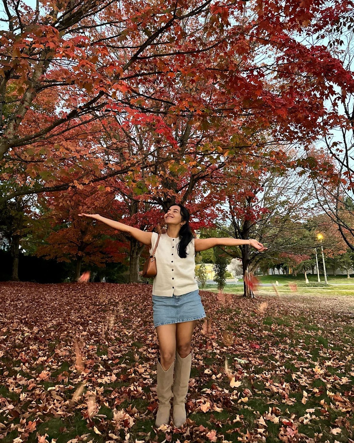 A woman in a denim skirt and tall boots stands under red autumn trees.