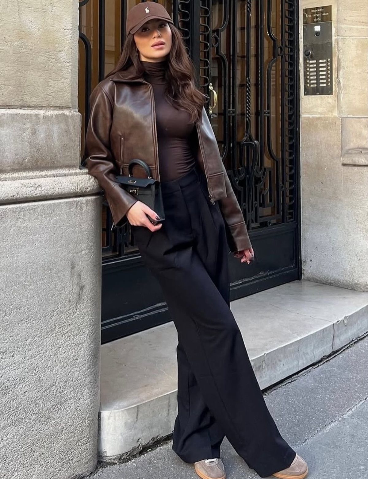 Woman in a brown leather jacket and black trousers leaning against a stone wall.