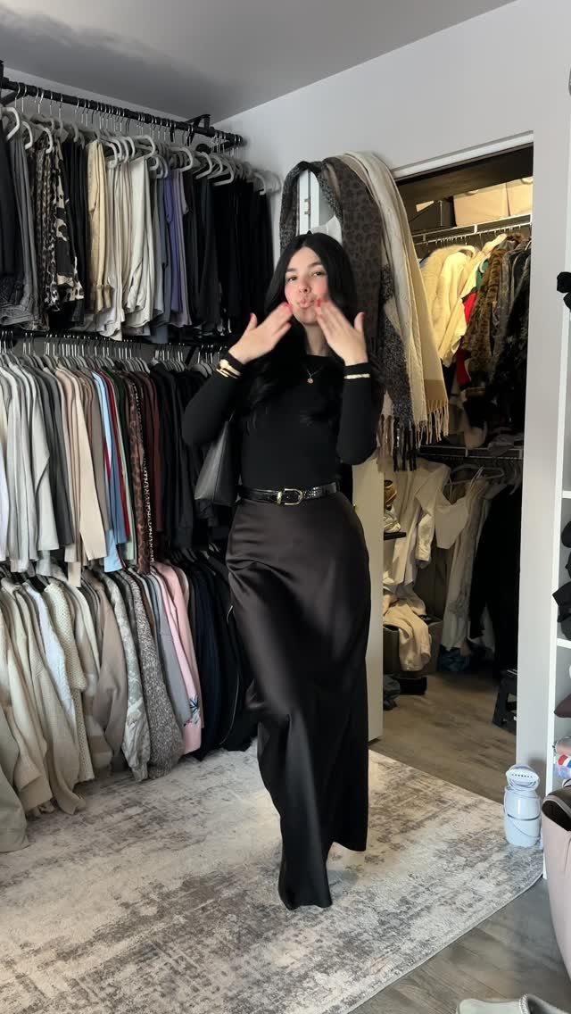 A woman posing in a black long-sleeved top and a long black satin skirt.