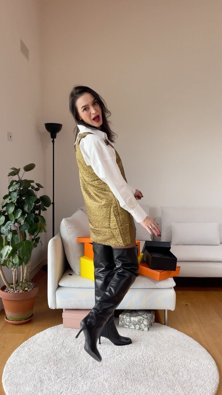 Woman wearing a gold mini dress and black leather over-the-knee high-heeled boots posing indoors.