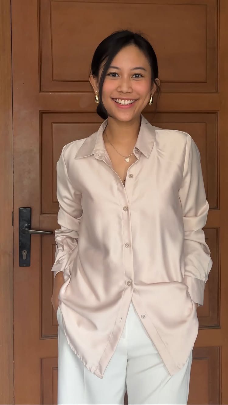 A smiling woman wearing a beige satin blouse and white pants standing before a wooden door.