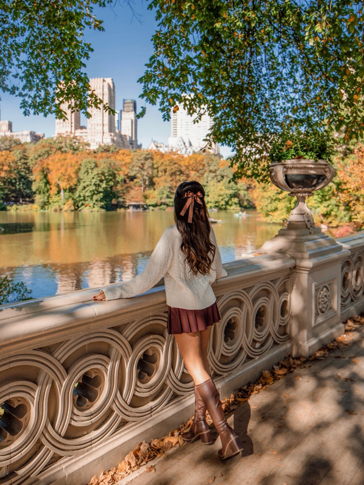 Woman in a white sweater, dark skirt, and brown boots standing by a lake in autumn.