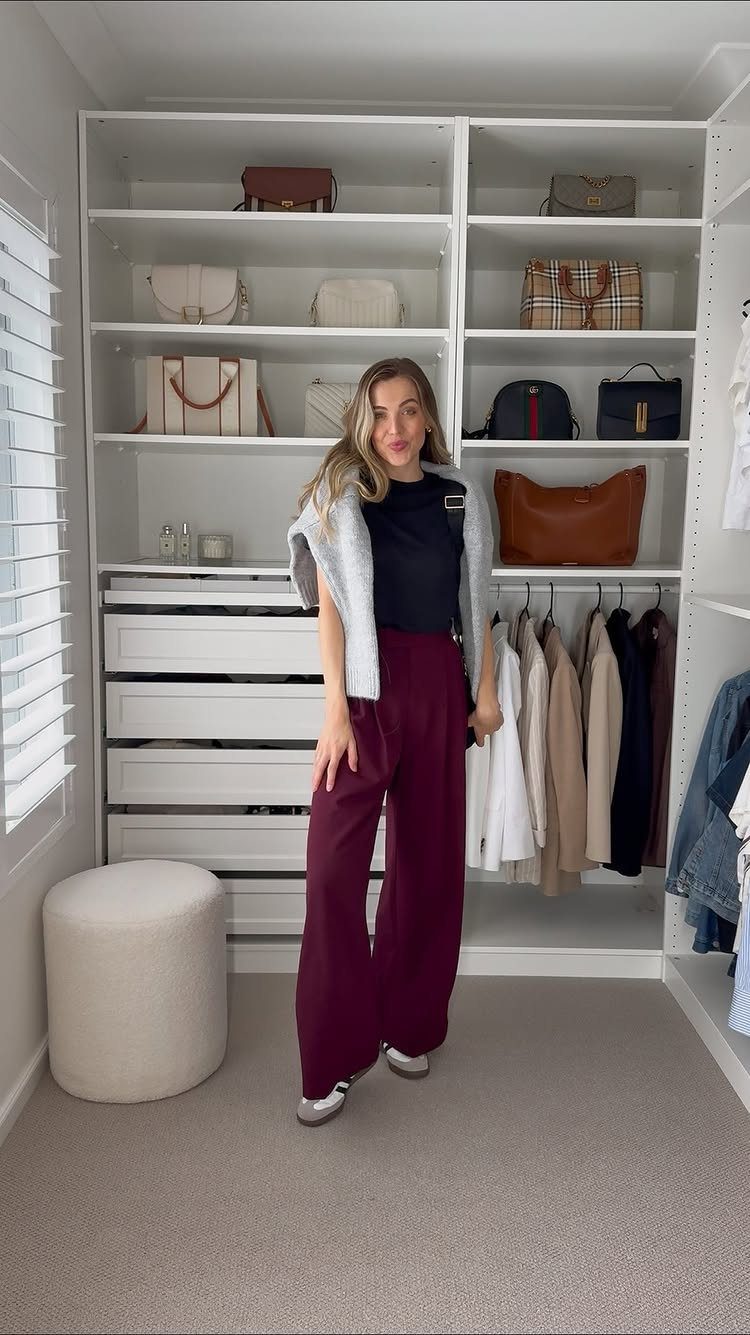 A woman posing in a closet wearing black top, grey cardigan, and burgundy wide-leg tailored pants.