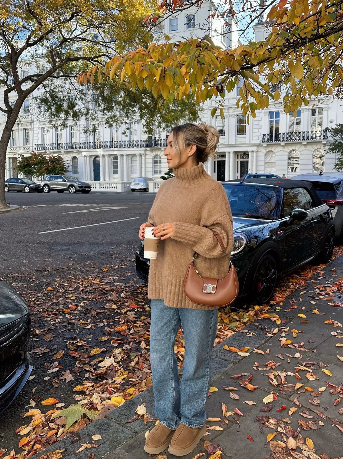 Woman in a tan turtleneck sweater and blue jeans standing on a leaf-covered sidewalk.