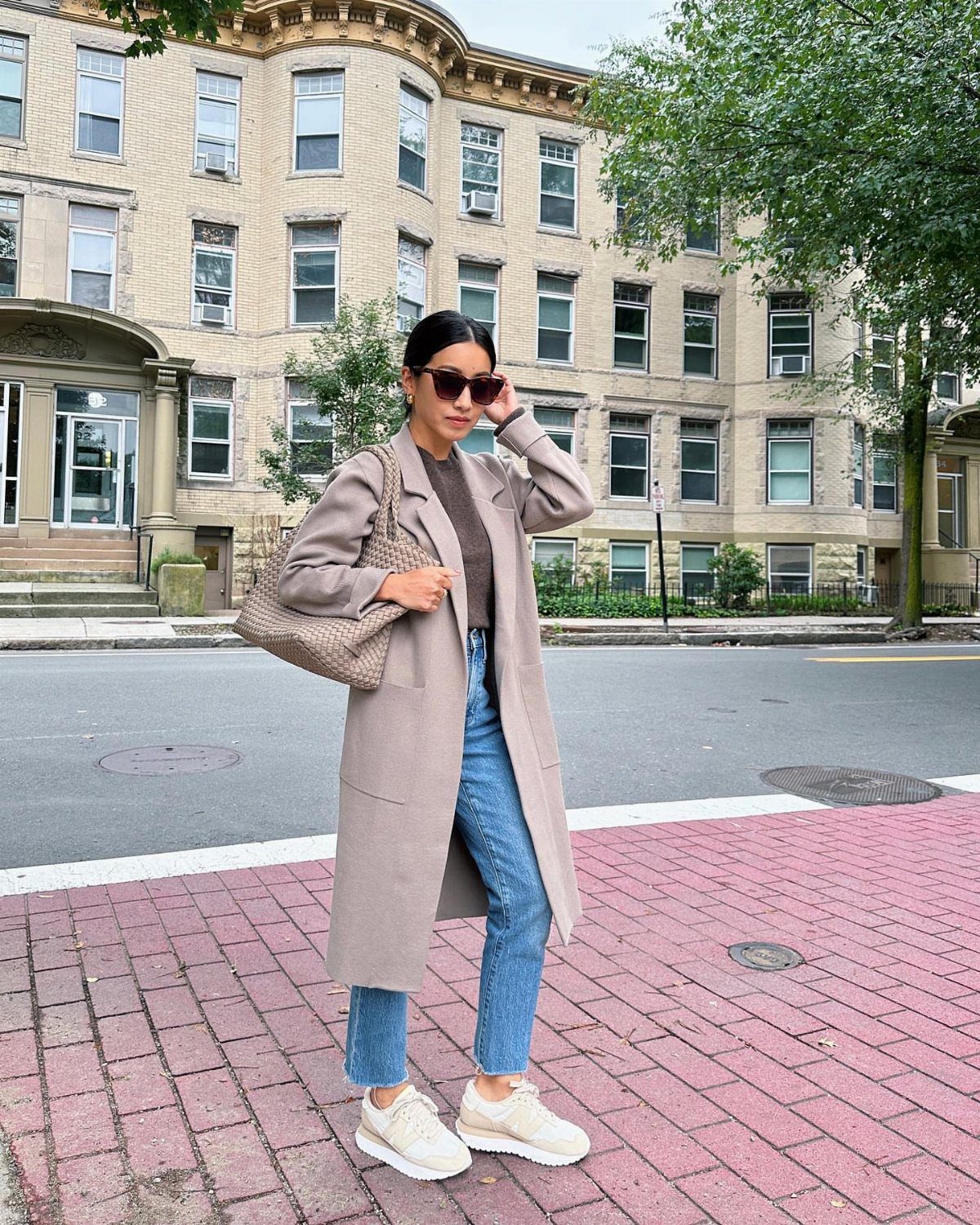 A woman wearing a long beige coat, blue jeans, and white sneakers standing on a city sidewalk.