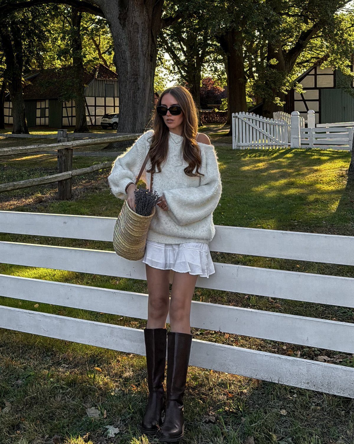 Woman in a white chunky knit sweater and ruffled skirt standing by a white fence outdoors.