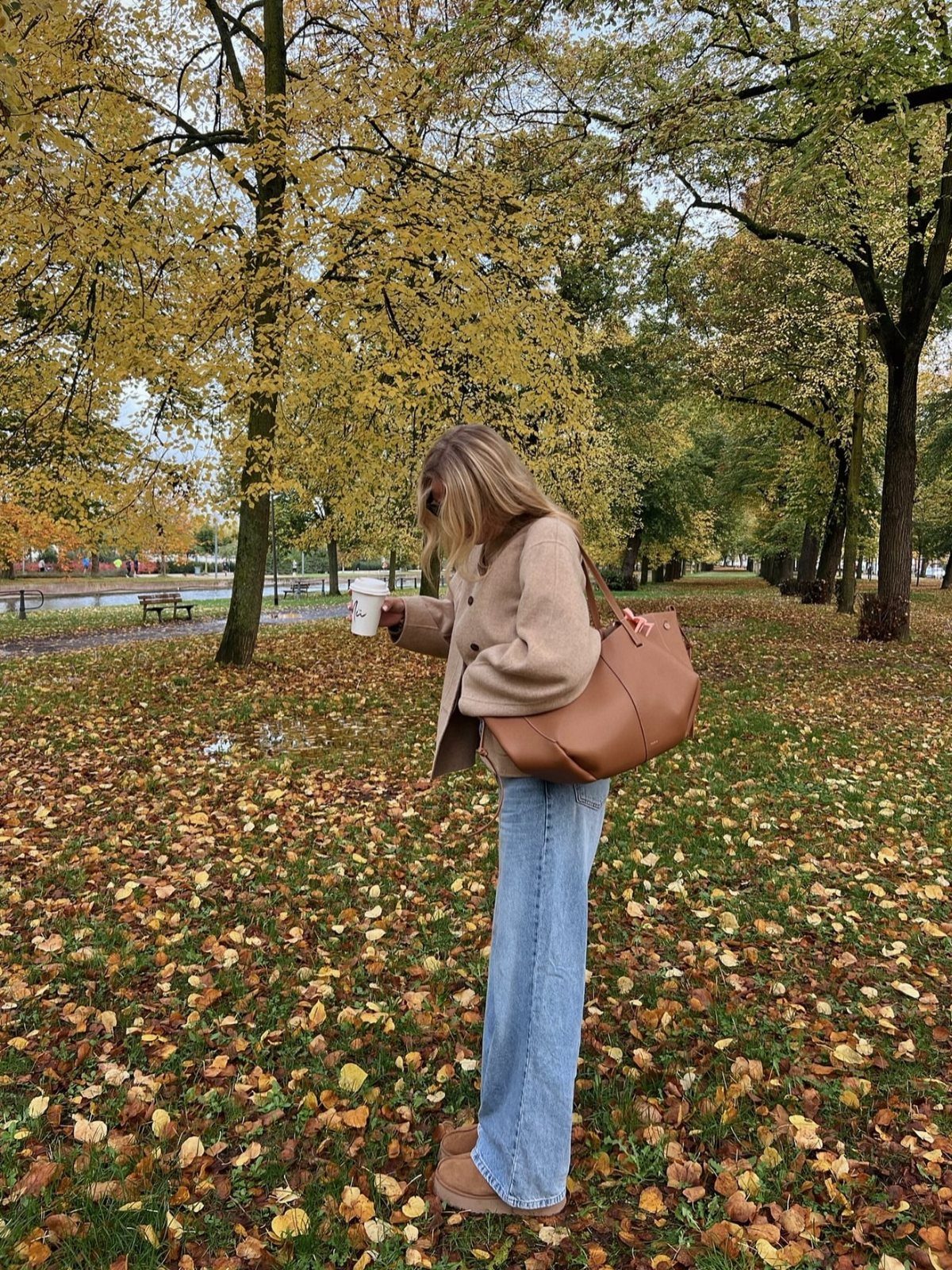 Woman in a tan blazer and light-wash denim jeans walking through a park with autumn leaves.