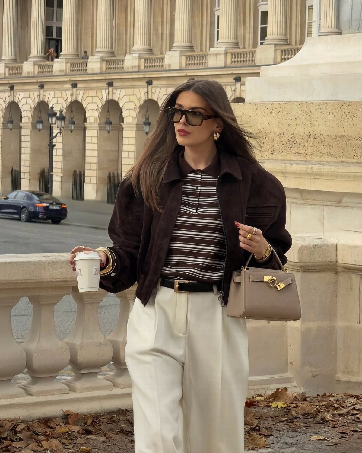 A woman in a brown jacket and cream trousers standing near a classical building.