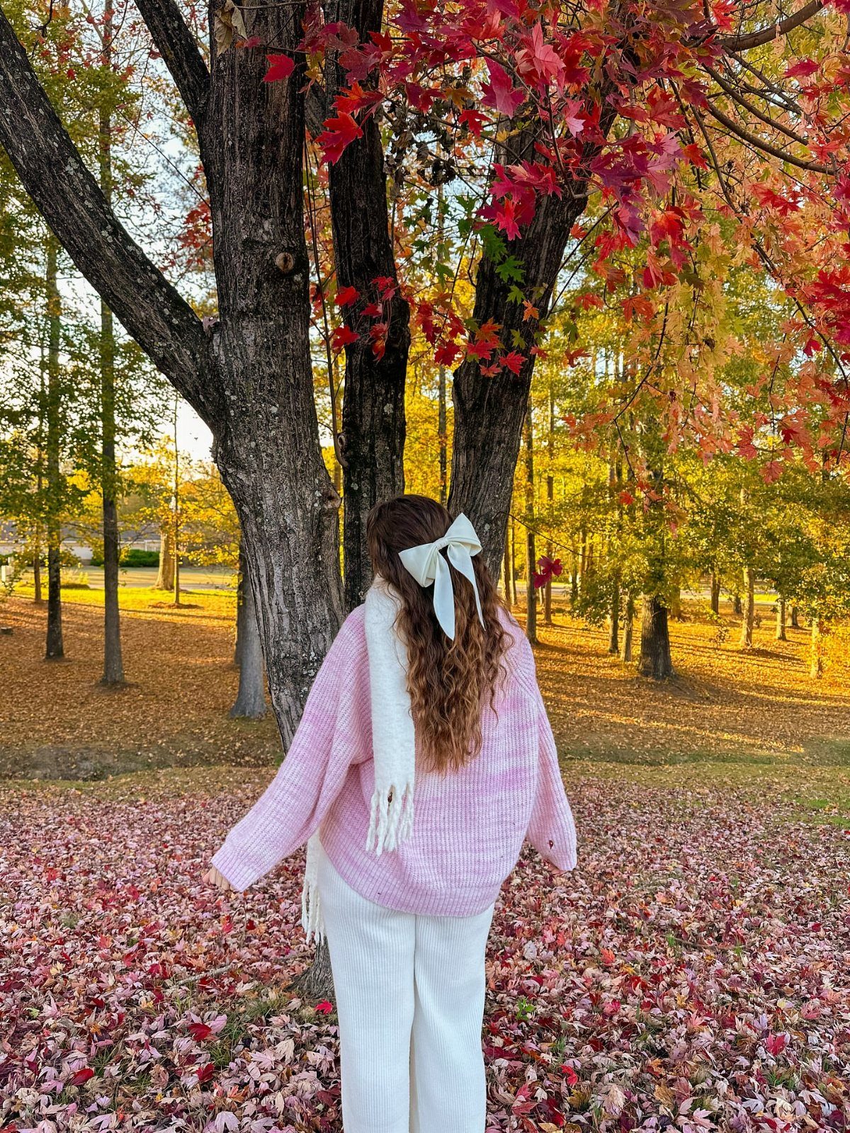 Woman in a pink sweater and white trousers standing among autumn trees and fallen leaves.