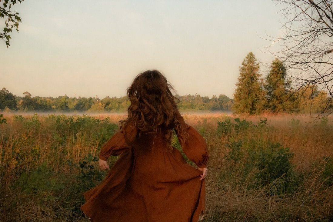 A woman in a rust-colored dress standing in a misty autumn field at sunset.