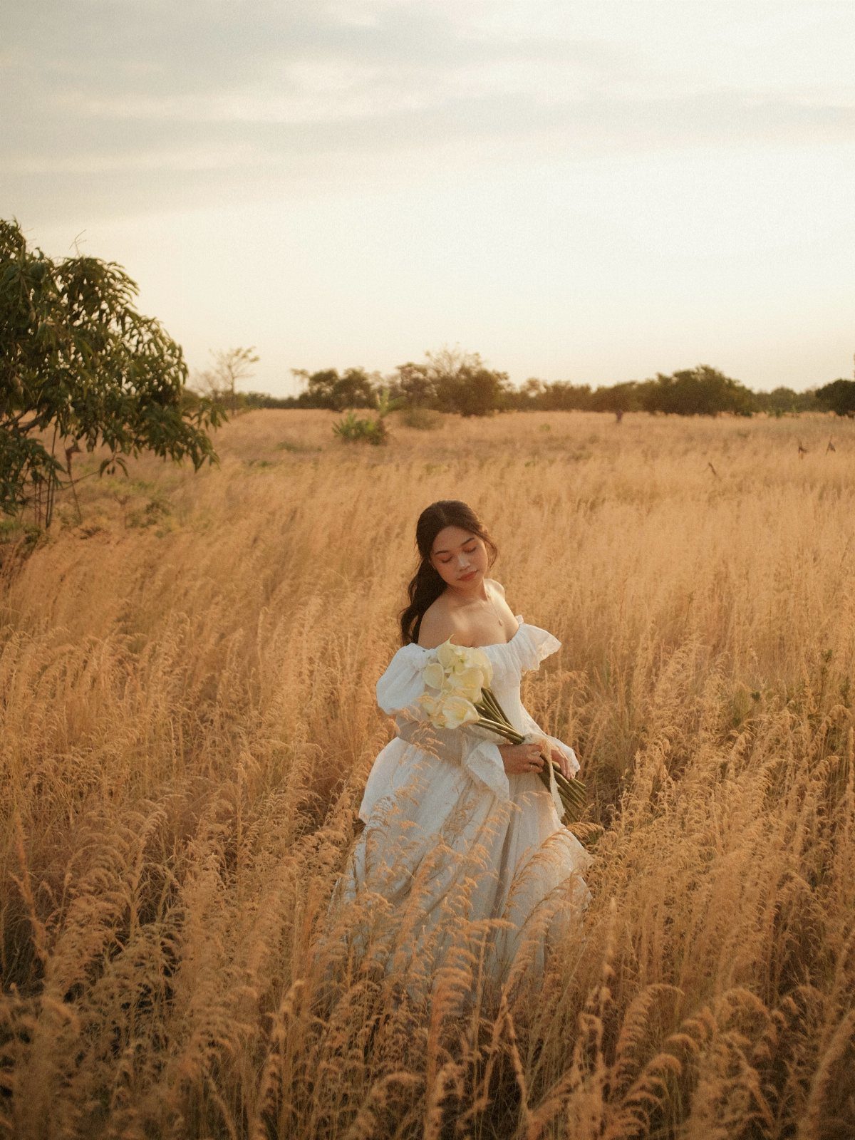 A woman wearing a white maxi dress holding flowers in a golden field.