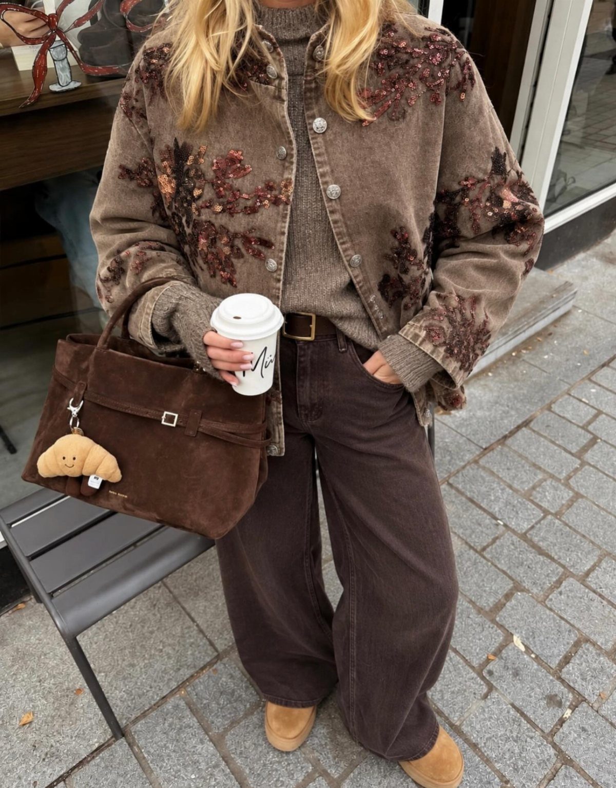 Woman wearing an embroidered brown corduroy jacket, tan sweater, and wide-leg brown trousers holding coffee.