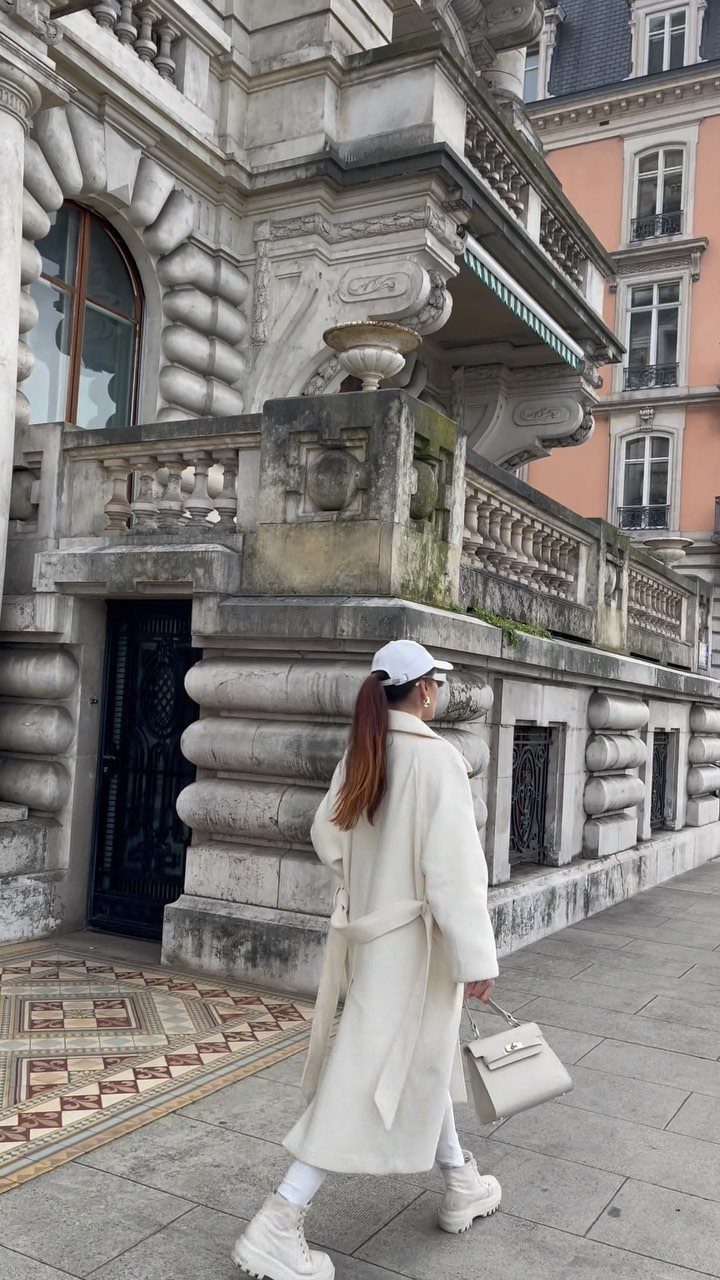 Woman walking in an all-white monochrome outfit past a large, ornate stone building.