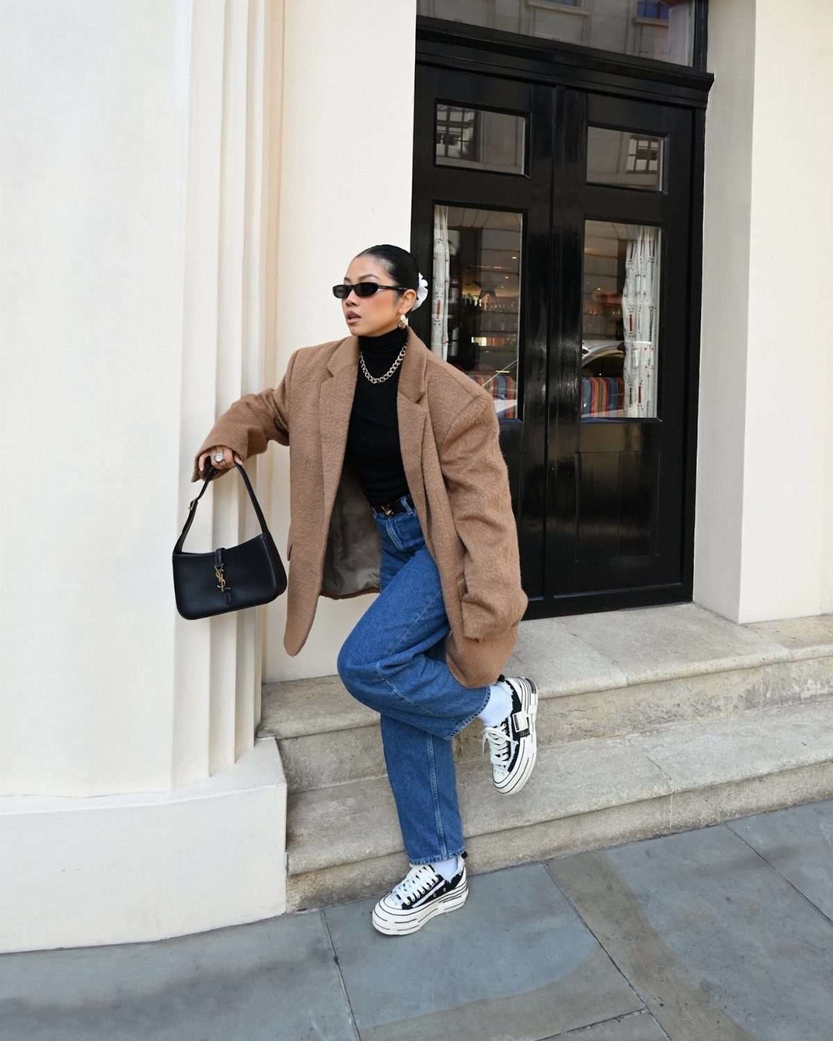 Woman in a brown oversized blazer, black turtleneck, and blue jeans standing on stone steps.
